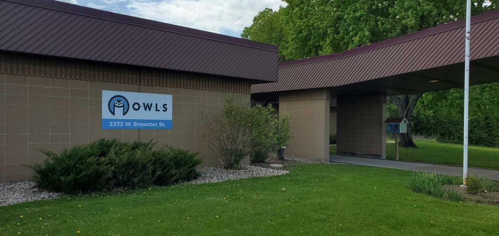 A beige brick building with a brown roof, a blue sign, and green lawn with bushes.