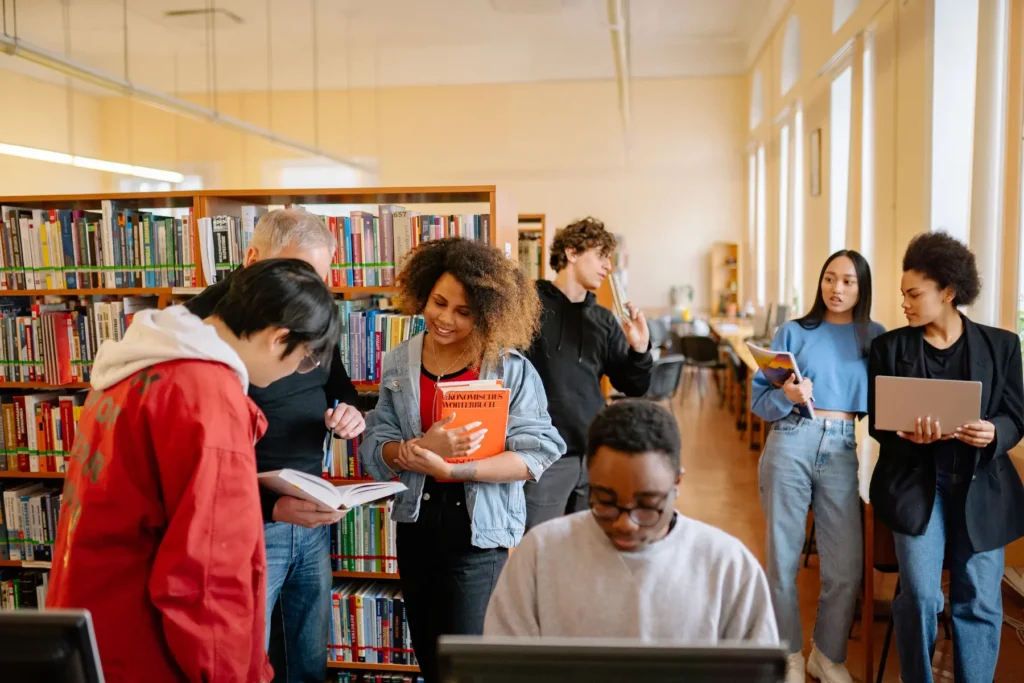 A diverse group of people interacting in a library, with bookshelves and large windows in the background.