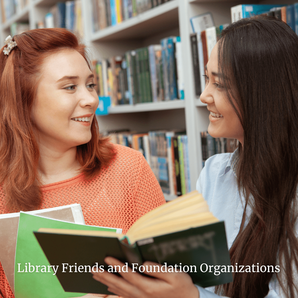 Two young women smiling and discussing books in a library, with bookshelves filled with various books behind them.